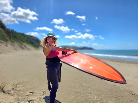 Cheerful woman on a sandy beach giving a peace sign, holding her surfboard, dressed in Tamara swimwear and wetsuit, ready for an empowering surf adventure.