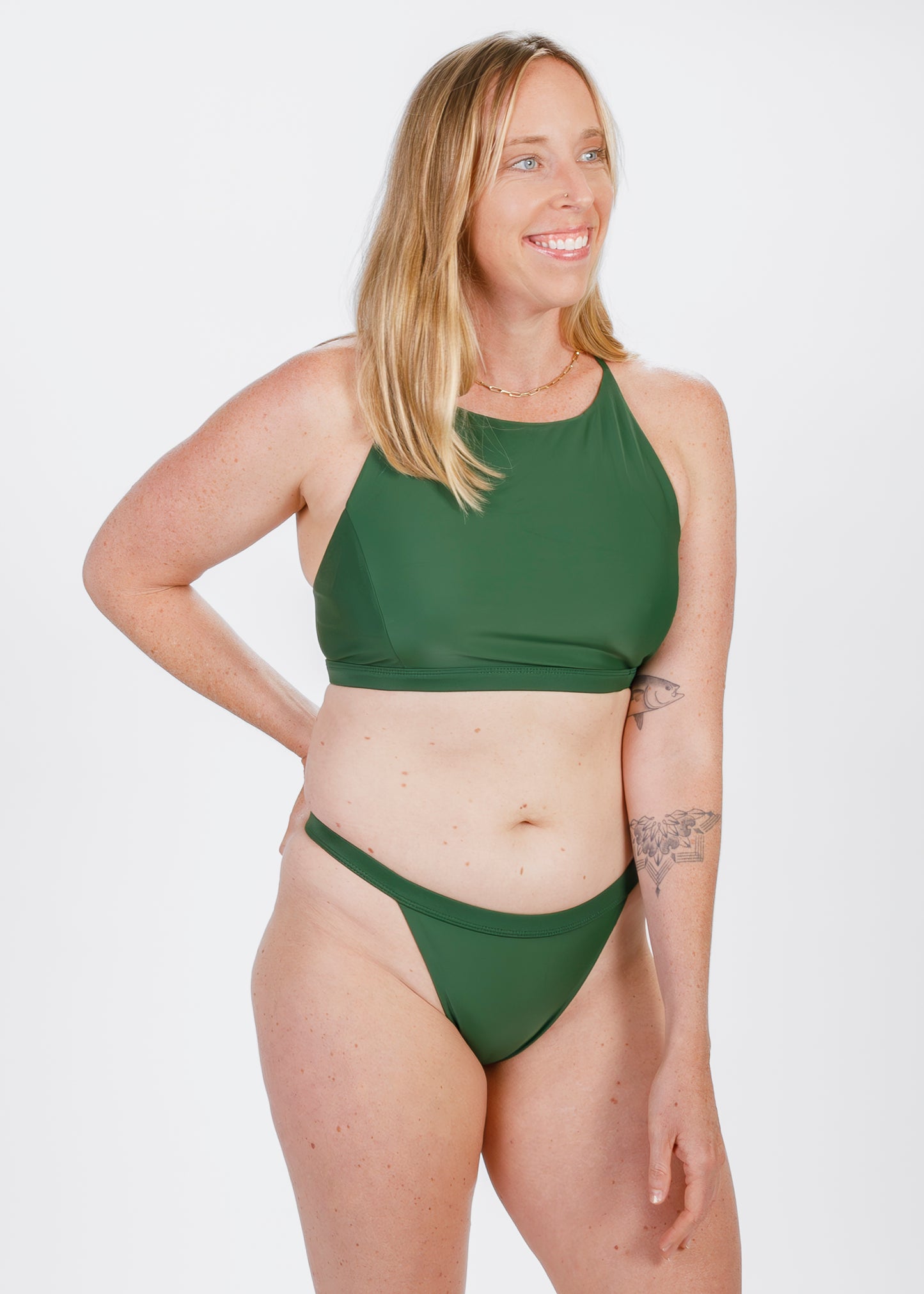 Model smiling and adjusting the waistband of her green bikini bottoms, exuding confidence in a studio setting.