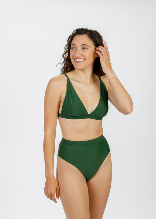 Model smiling and adjusting her green high-waisted bikini, while lightly touching her hair in a relaxed pose.