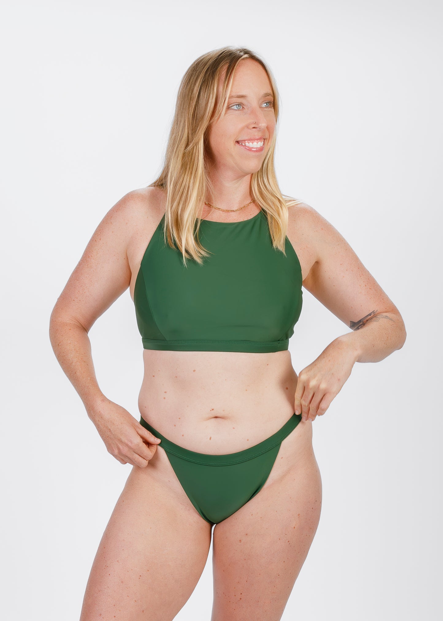 Model smiling and adjusting the waistband of her green high-waisted bikini, captured in a studio with a relaxed, confident expression.