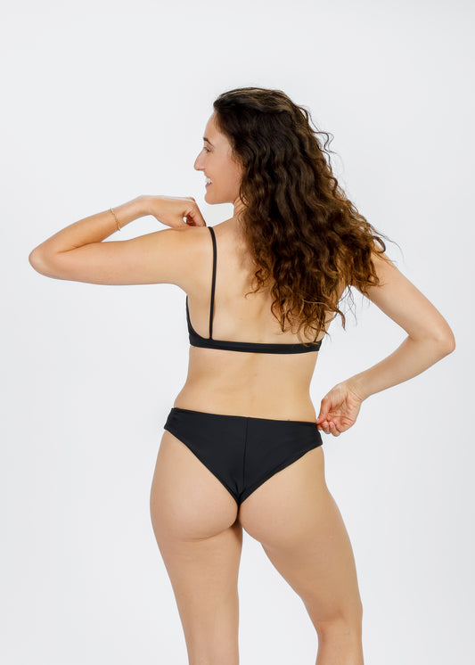 Woman with curly hair wearing a black two-piece bikini, shown from behind as she adjusts her bottoms and smiles over her shoulder.