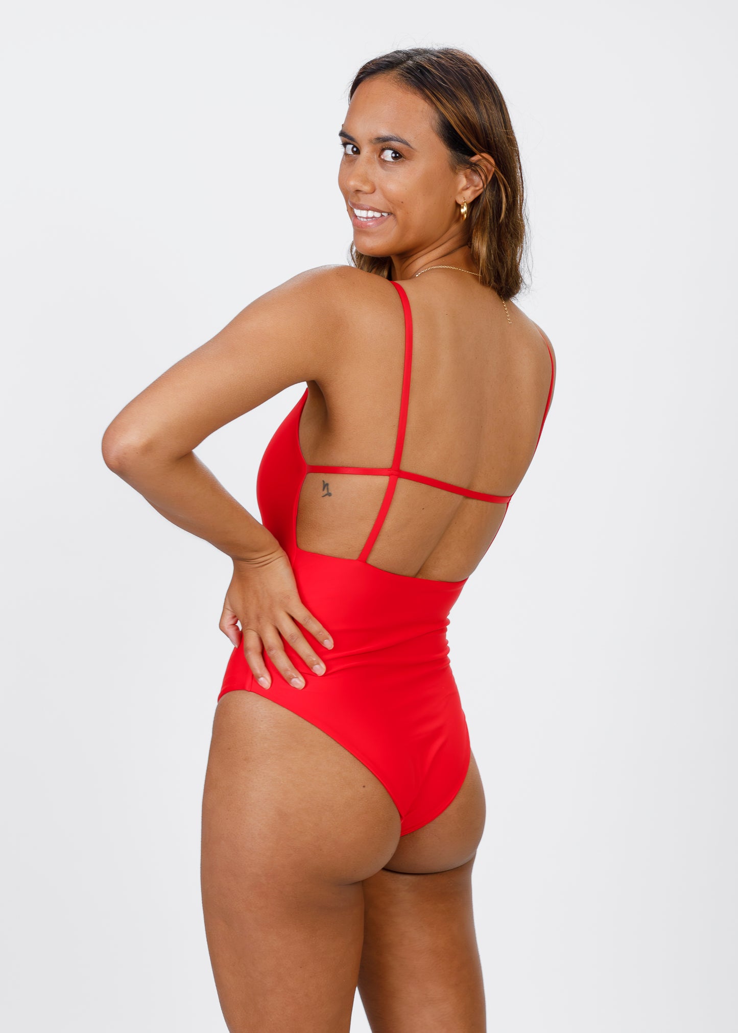 Woman turning to smile at the camera in a red, thin-strapped one-piece swimsuit with an open back, hand resting near her hip.
