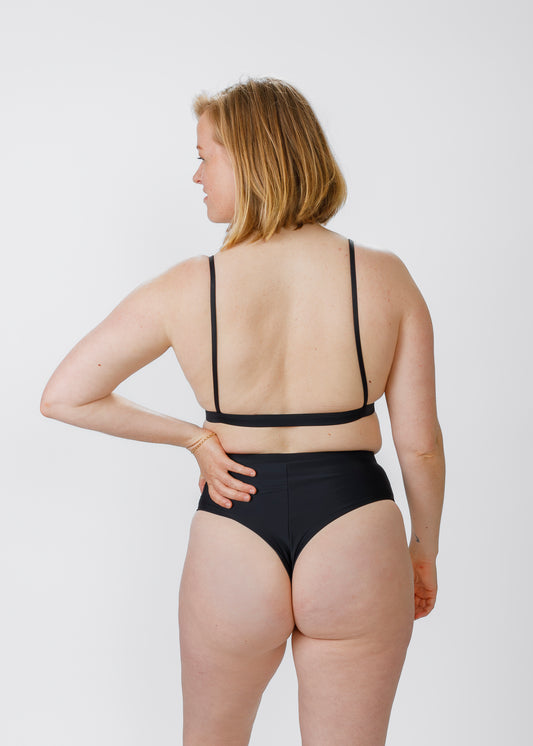 A blonde woman stands with her back to the camera, wearing a thin-strap black bikini top and matching high-waisted bottoms. She rests one hand on her hip, emphasizing the secure, flattering cut of the bottoms.