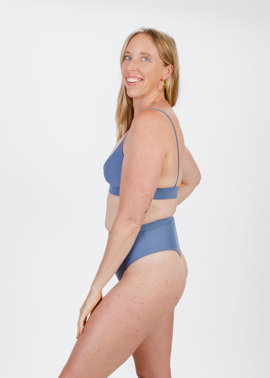 A woman is shown from the back, wearing a matching bikini top and high-waisted bottoms. She places one hand on her hip, emphasizing the waistband’s secure fit and flattering coverage.