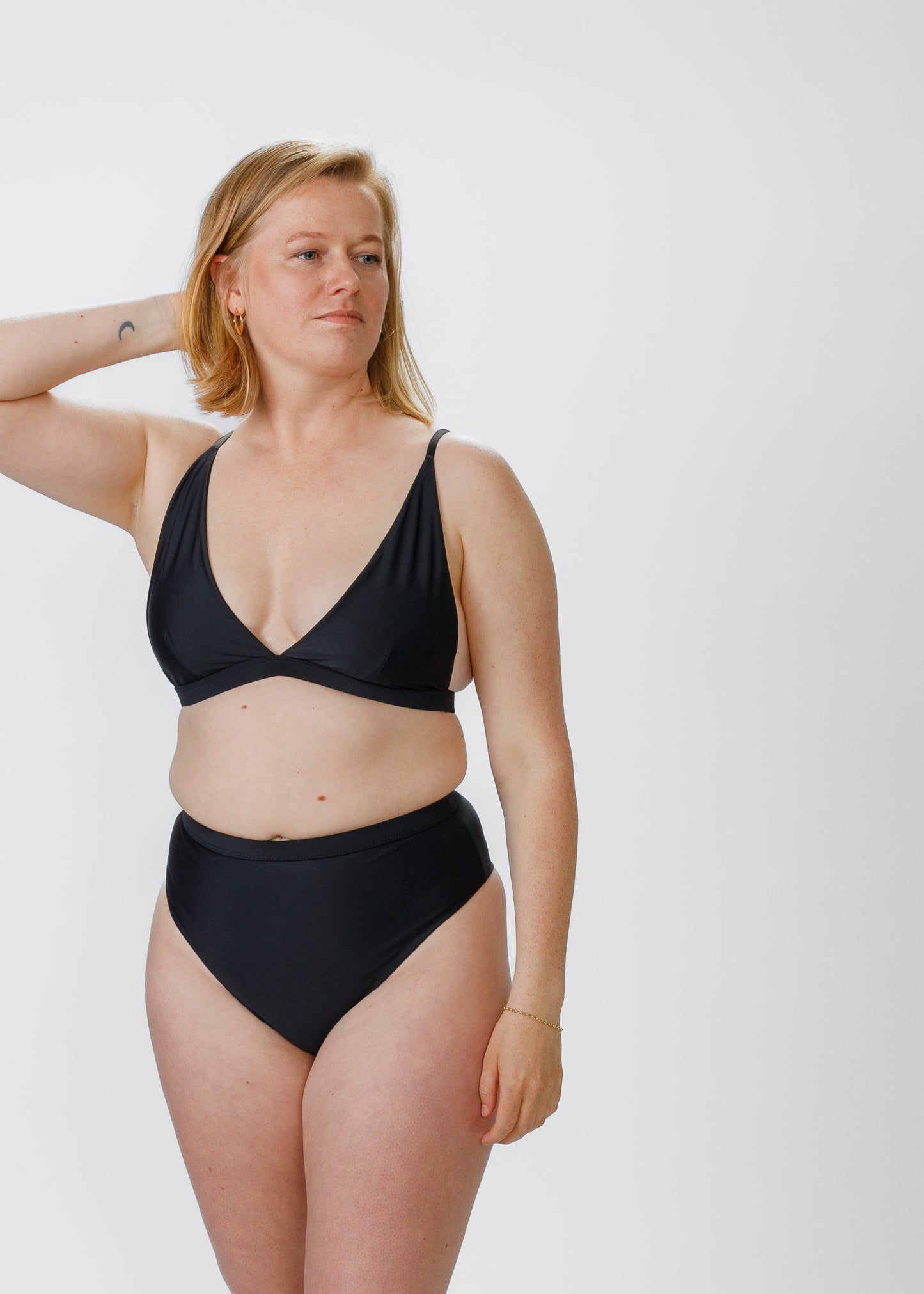 Model wearing a black high-waisted bikini, with one hand resting on her neck and a relaxed pose, looking away from the camera.