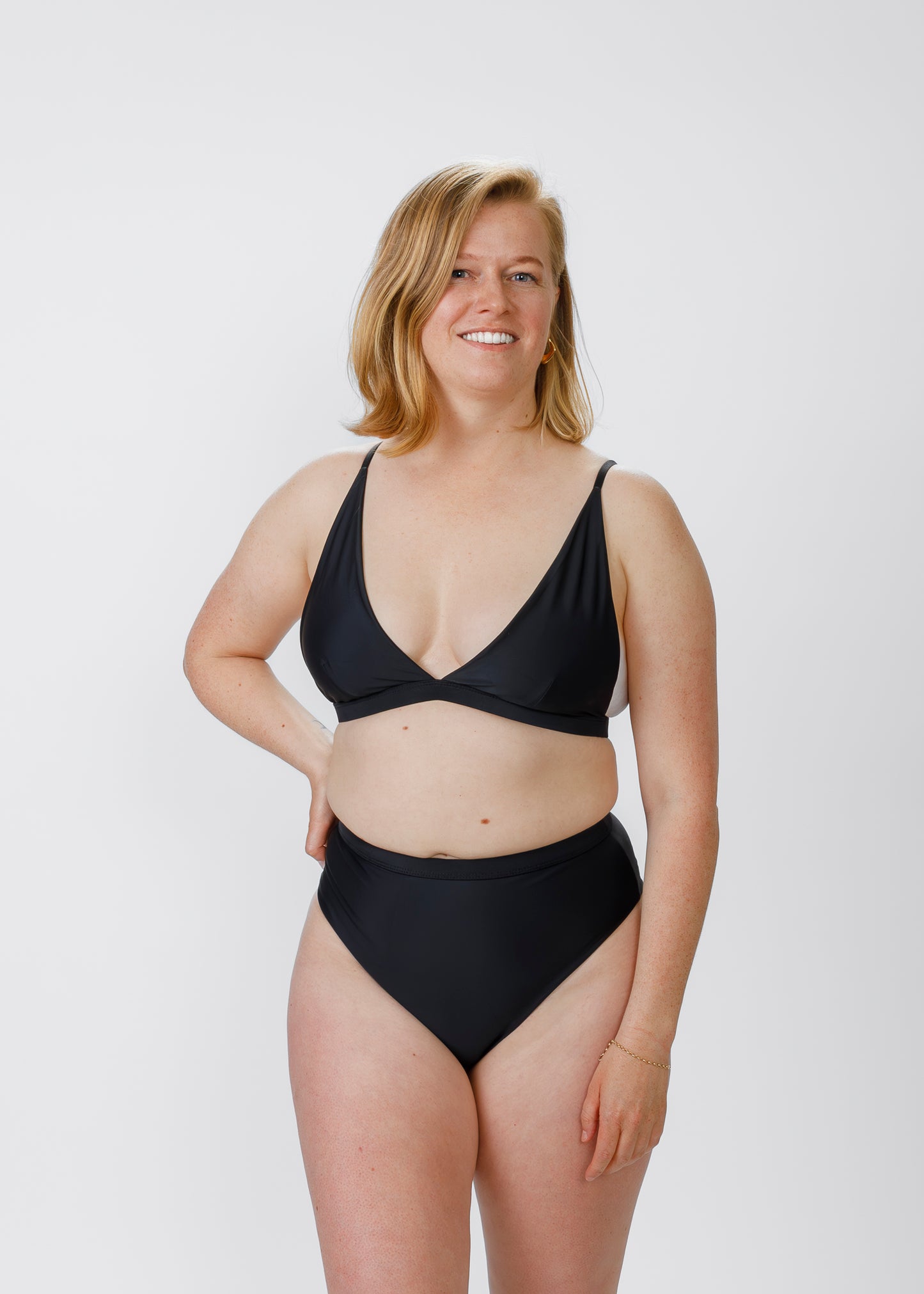 Model wearing a black high-waisted bikini, smiling with a hand on her hip, facing the camera in a studio setting.
