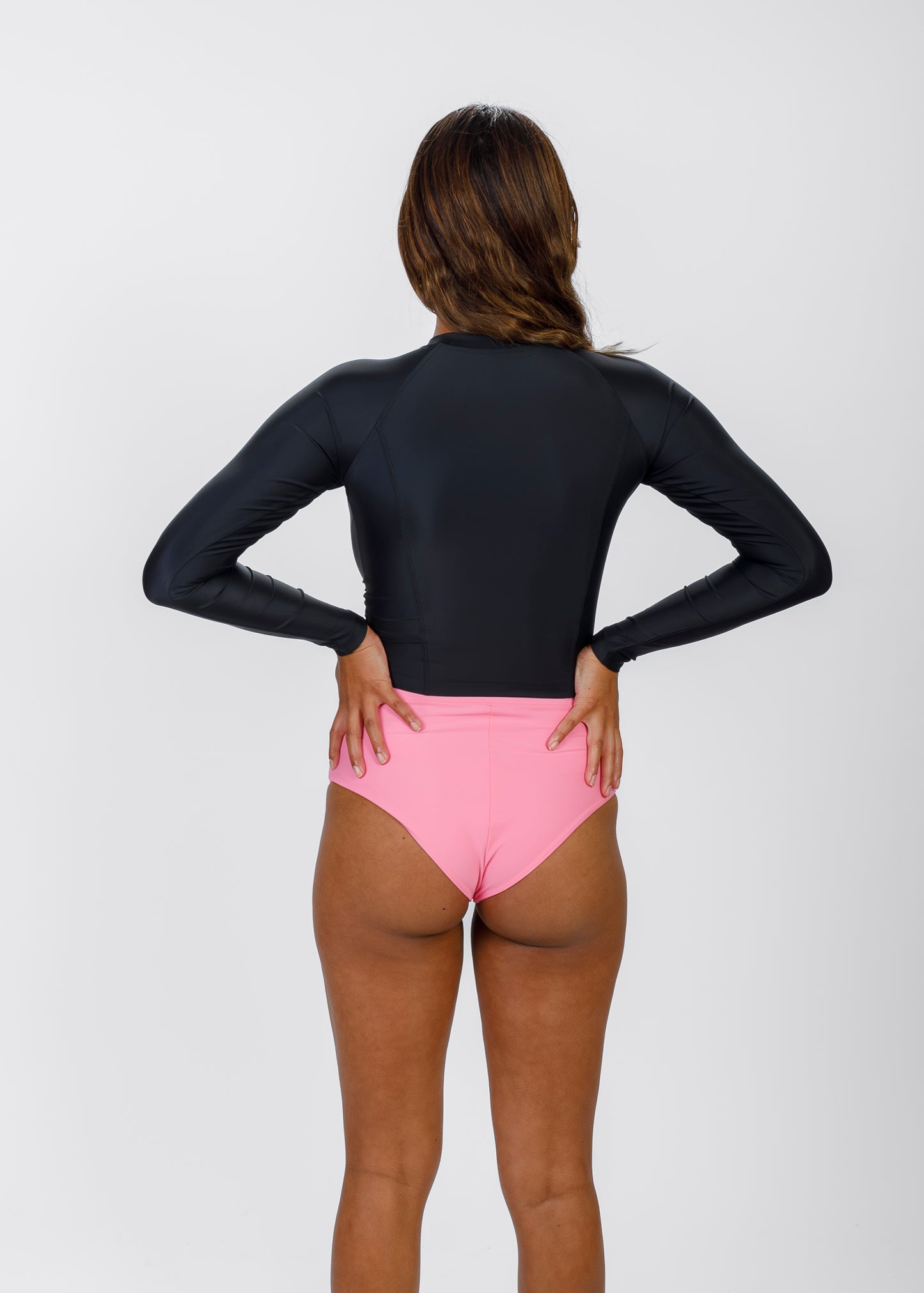 Back view of a woman in a black Tamara rash guard and pink high-waisted bikini bottoms, hands on her hips, facing away from the camera.