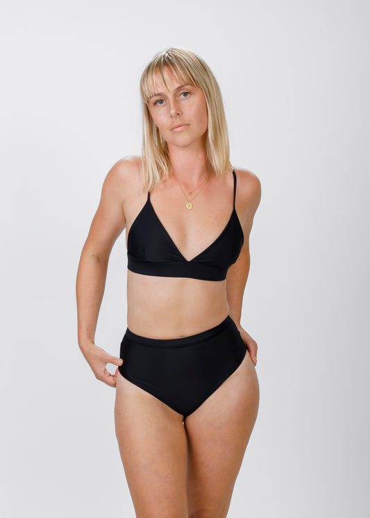 A blonde woman faces forward, wearing a black triangle bikini top with a subtle V-neck. She lightly touches the high-waisted bottoms, emphasizing the top’s flattering cut and the brand’s focus on supportive, timeless swimwear.