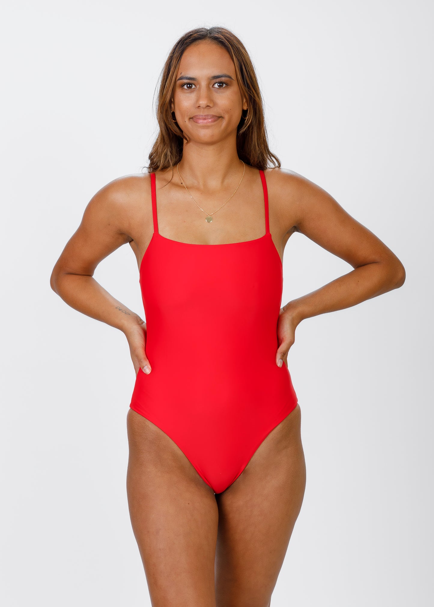 Woman wearing a bright red, thin-strapped swimsuit with a square neckline, standing with hands on her hips and looking straight at the camera.