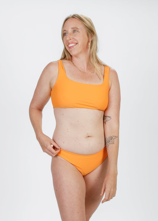 Smiling blonde woman wearing a bright orange, square-neck bikini top and matching bottoms, adjusting the waistband while looking off to the side.