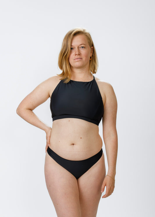 A fair-skinned, blonde woman stands against a white backdrop wearing a black halter-style bikini top and matching bottoms. She has one hand on her hip and looks confidently at the camera, highlighting the swimsuit’s high neckline and supportive fit.