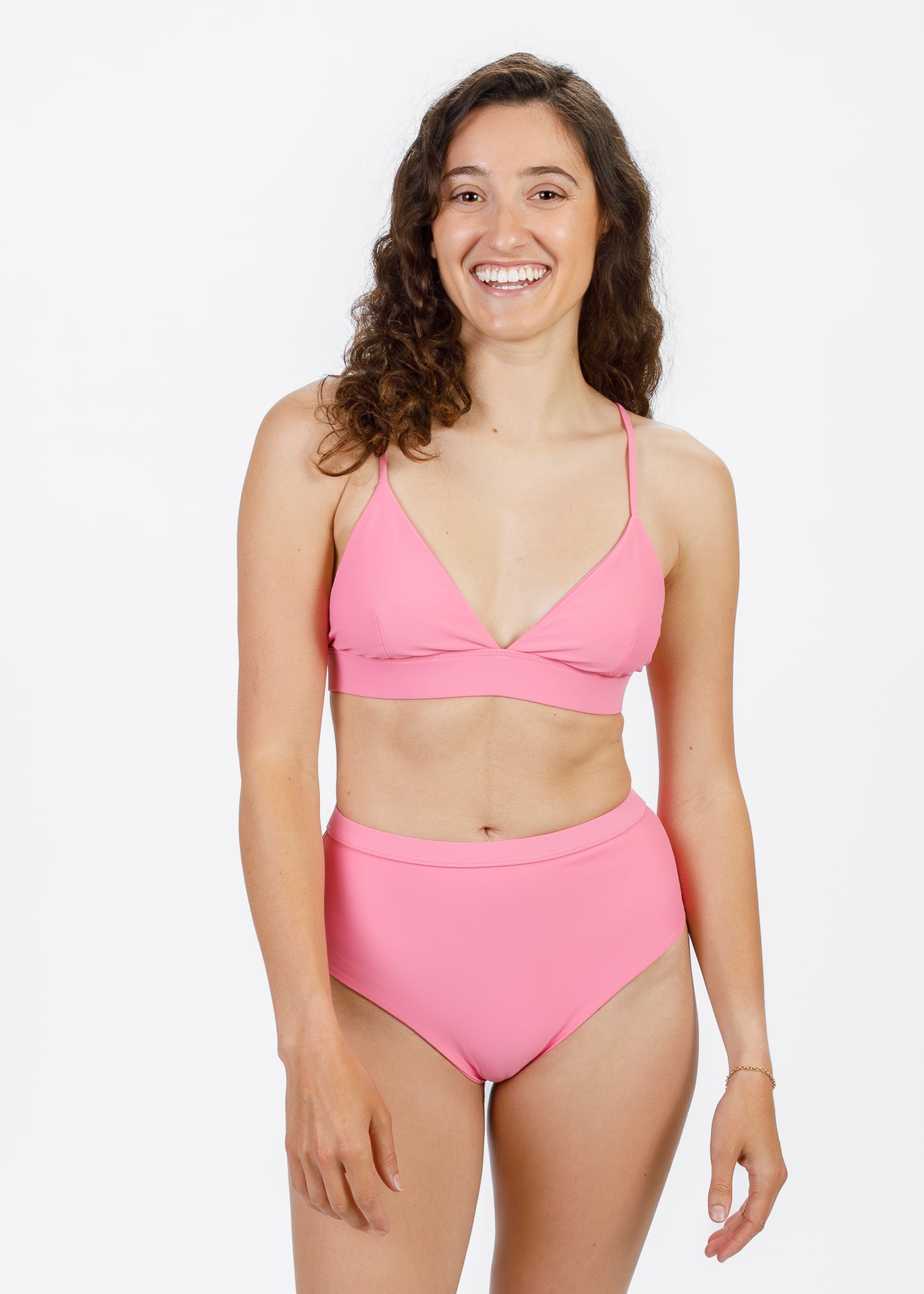 A brunette woman with curly hair stands facing forward in a pink triangle bikini top with a wide underband. She smiles brightly, showcasing the top’s minimal coverage and adjustable straps for an easy, confident fit.