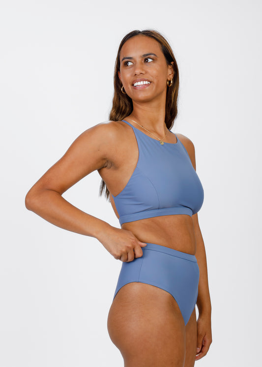 A woman in a light blue high-neck bikini adjusts the waistband of her matching high-waisted bottoms. This angle highlights the flattering cut and secure fit designed for confident, eco-friendly beachwear.