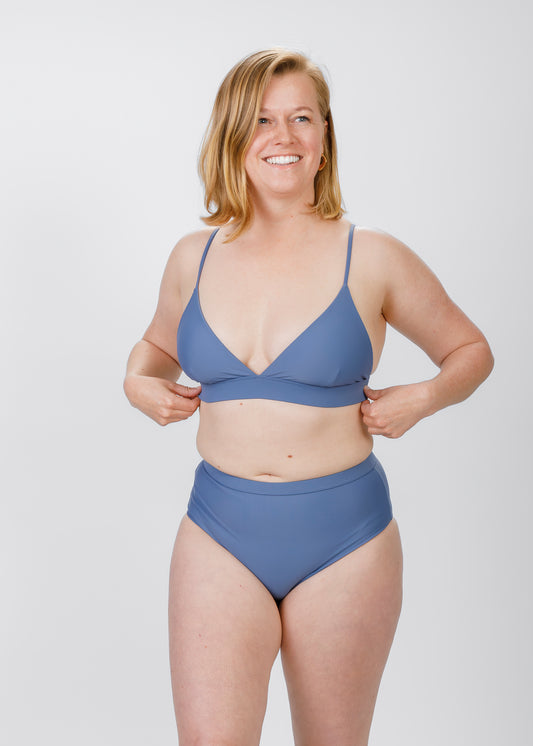 A fair-skinned woman with a blonde bob smiles as she adjusts the underband of her light blue triangle bikini top. The delicate straps and softly plunging neckline reflect the brand’s modern design and inclusive sizing.