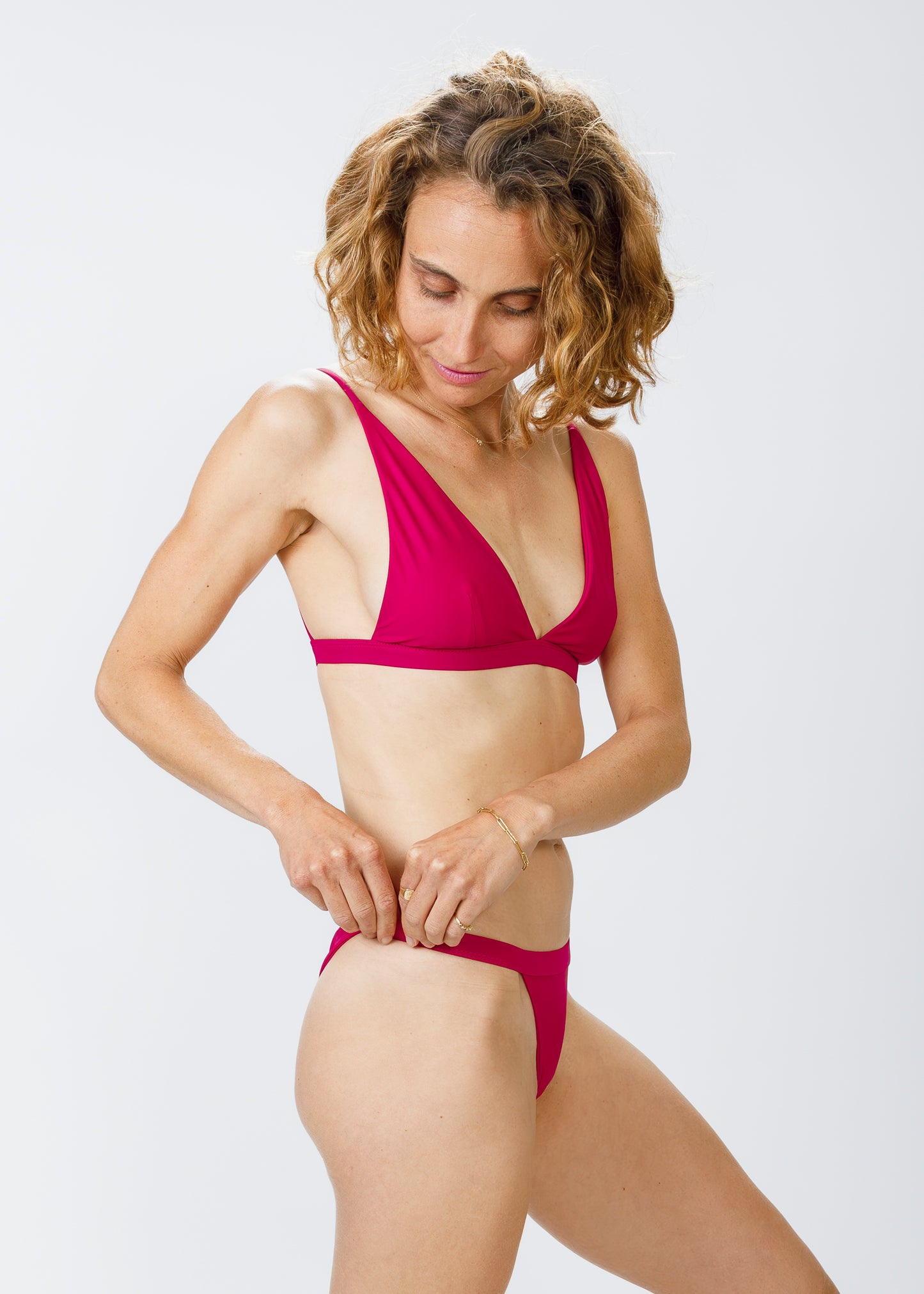 Model adjusting the side of her magenta bikini bottoms, wearing a matching bikini top, with a thoughtful expression.