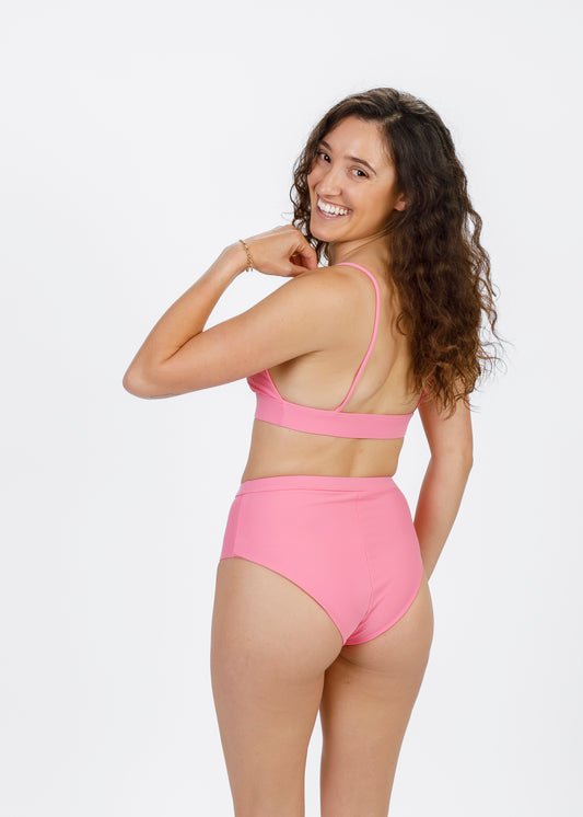 The same curly-haired woman turns to smile at the camera, revealing the thin straps and band of her pink triangle bikini top. Her high-waisted bottoms complete the playful, feminine look while emphasizing the top’s sleek, modern design.