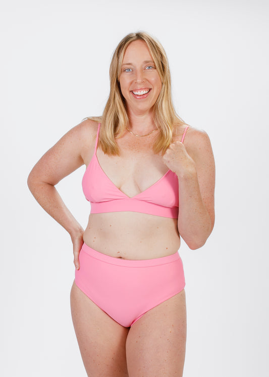 A blonde woman wearing a pink triangle bikini top with slender straps gently lifts one hand toward her shoulder. The soft, plunging neckline and supportive underband highlight the brand’s commitment to comfort and style.