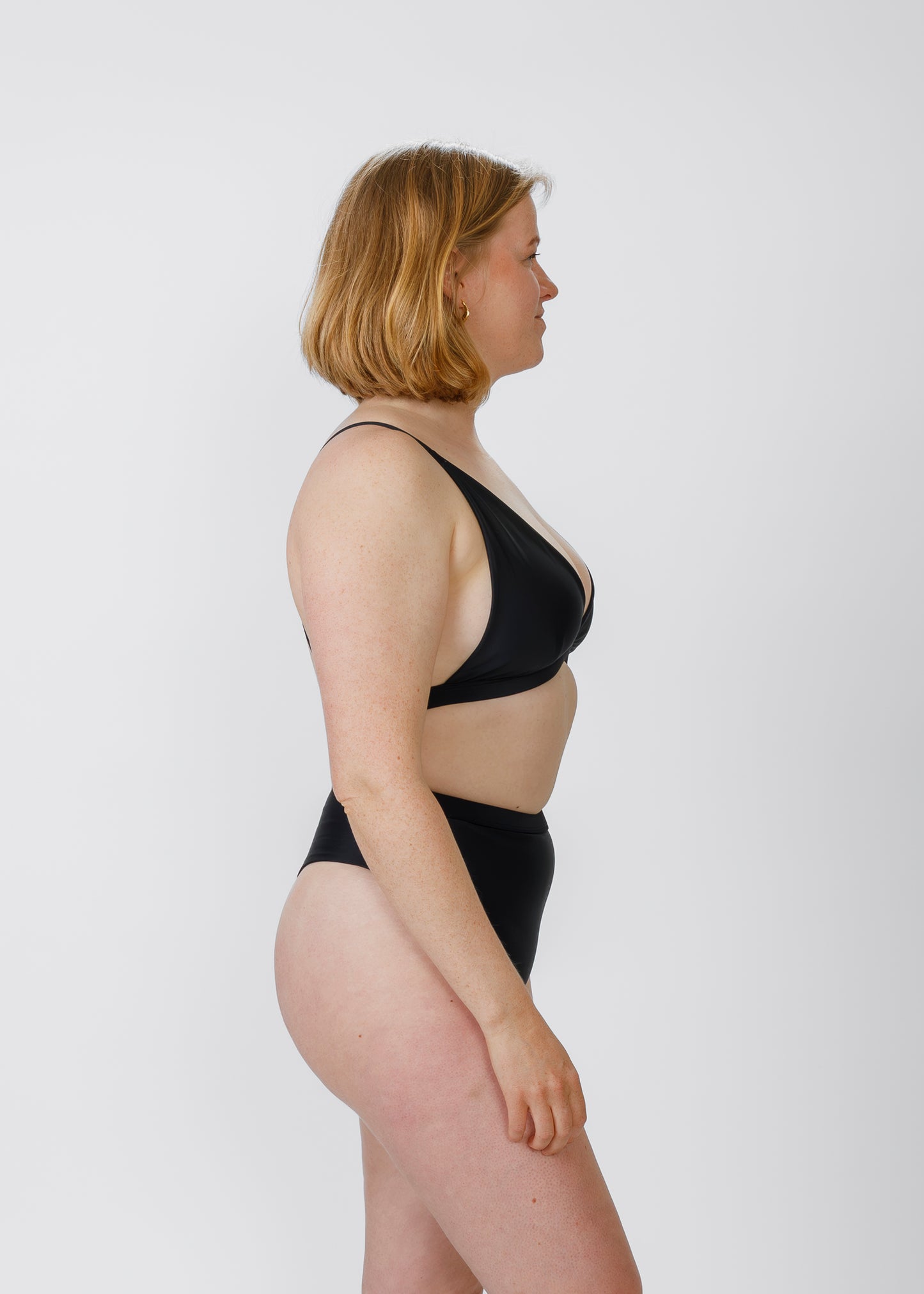 Side view of a model wearing a black high-waisted bikini, showing the profile of her figure with a neutral expression.