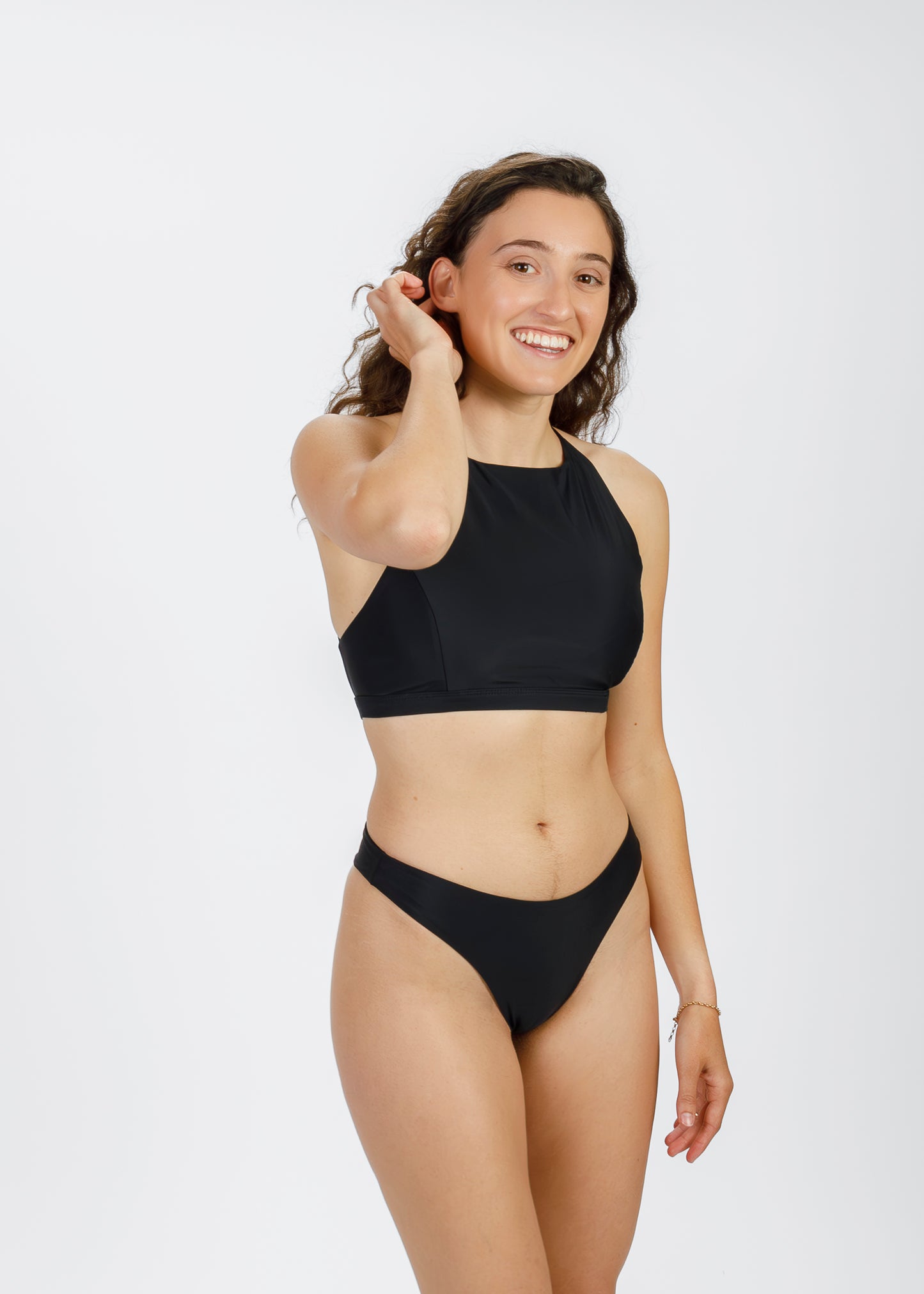 A woman with curly hair and a light-to-medium complexion stands in a black halter-style bikini, smiling and running her fingers through her hair. The modern, sustainable silhouette showcases the brand’s focus on comfort and versatility for all body types.
