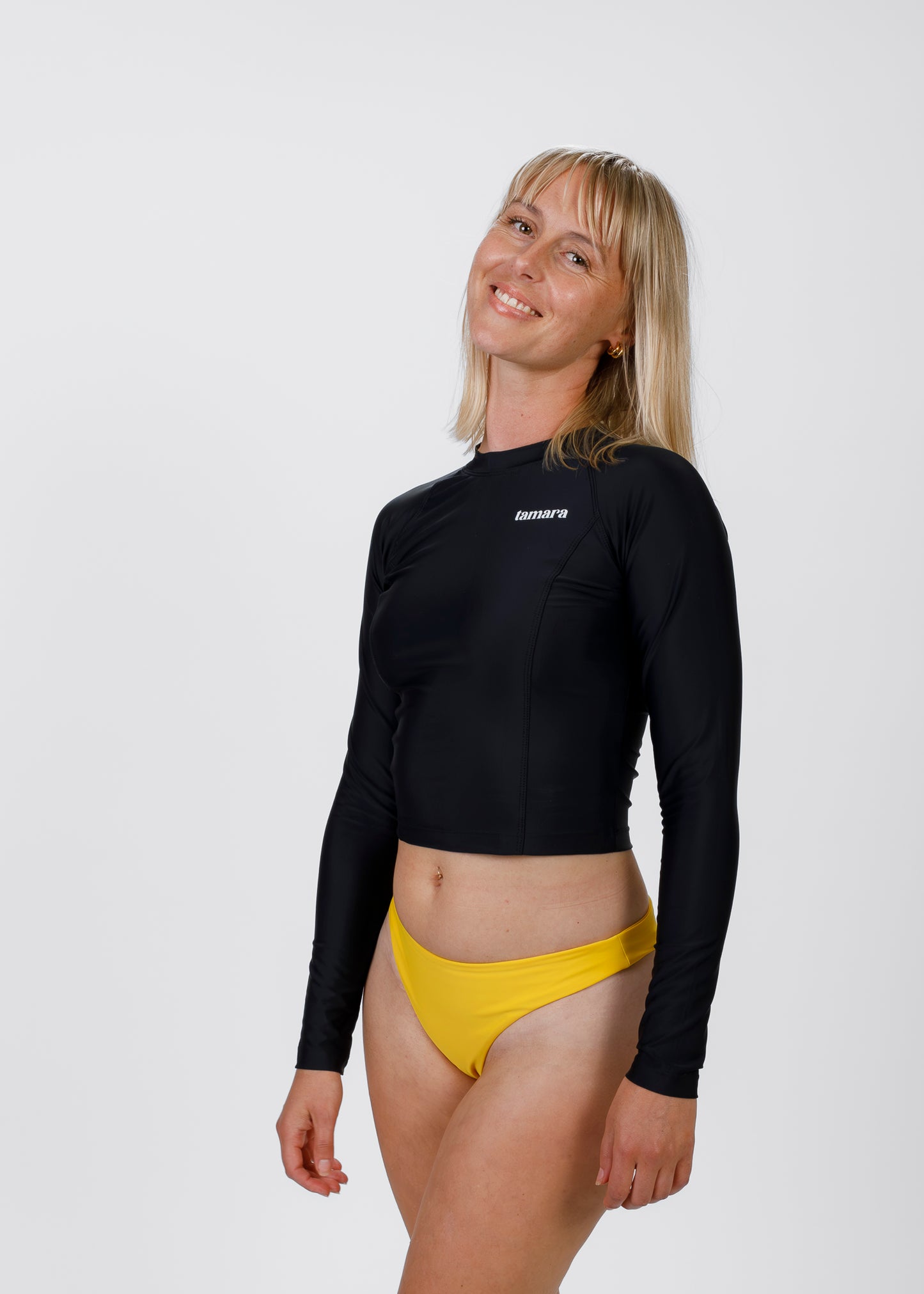 Blonde woman wearing Tamara’s black, long-sleeve crop rash guard and bright yellow bikini bottoms, slightly tilting her head and smiling against a white background.