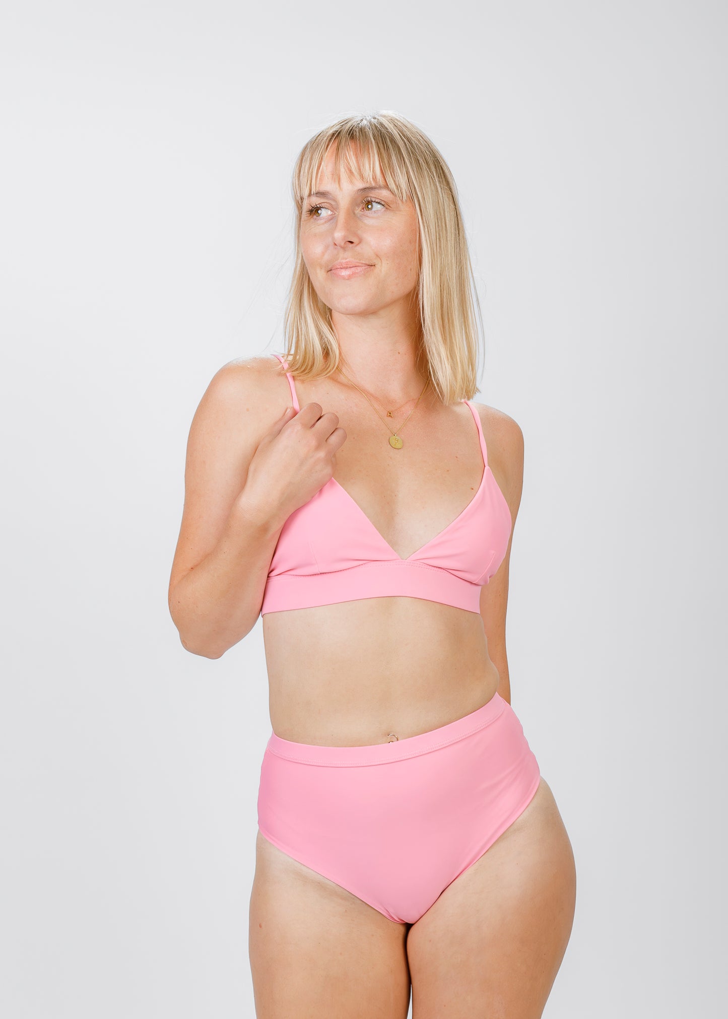A blonde woman gazes off-camera while wearing a pink triangle bikini top with slim straps and a supportive underband. She pairs it with high-waisted bottoms, highlighting the elegant, minimalist design of this eco-conscious swim set.