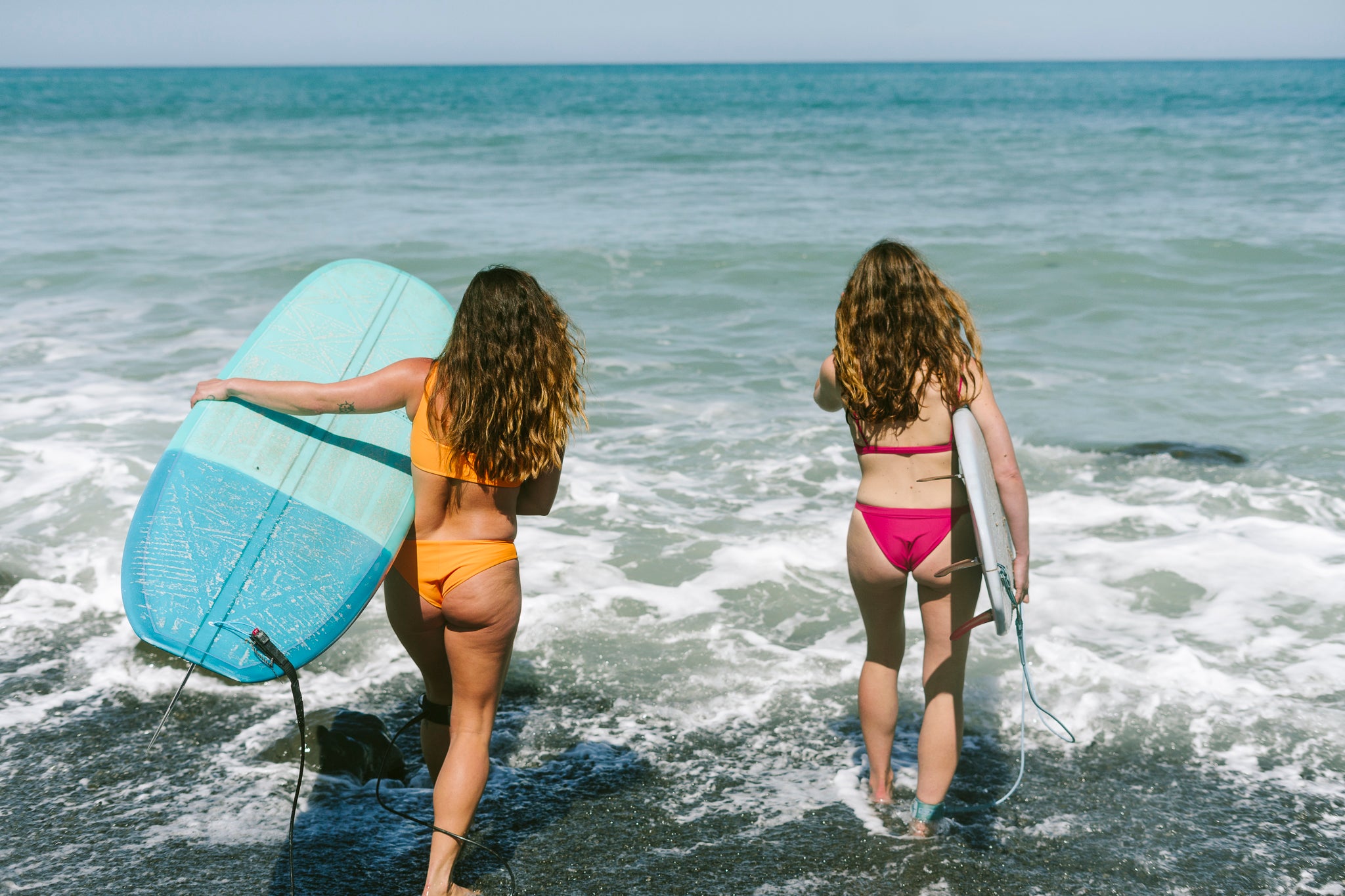 Two women walking into the ocean carrying surfboards, wearing bright orange and pink bikinis from Tamara™ swimwear, ready to surf.