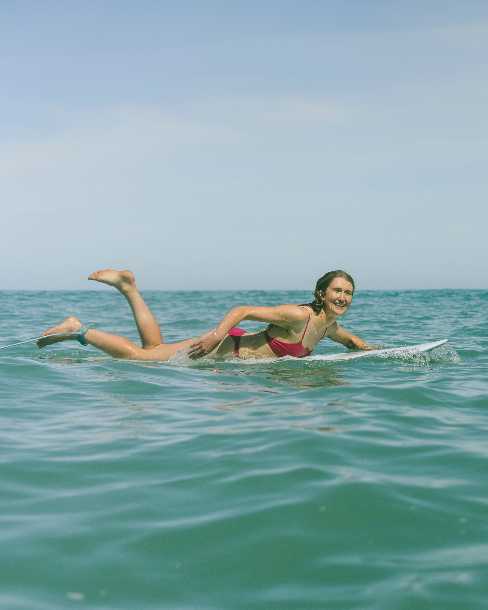 A woman wearing a dark pink bikini paddling on a surfboard in calm, open ocean waters under a clear blue sky.