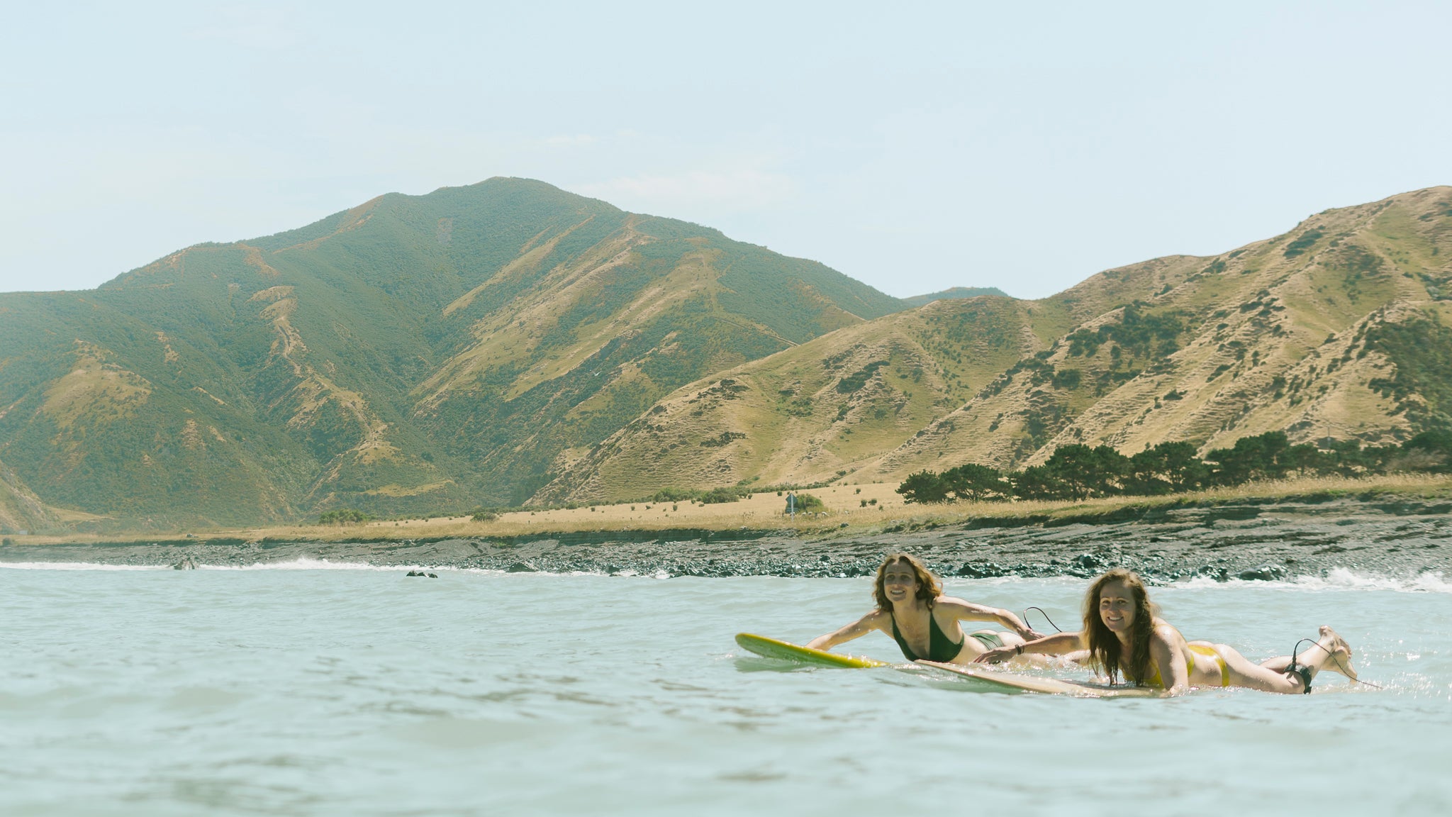 Two women lying on their surfboards, paddling through gentle ocean waves with lush, green hills and rocky shores in the background. They appear happy and relaxed, embracing the natural coastal environment.