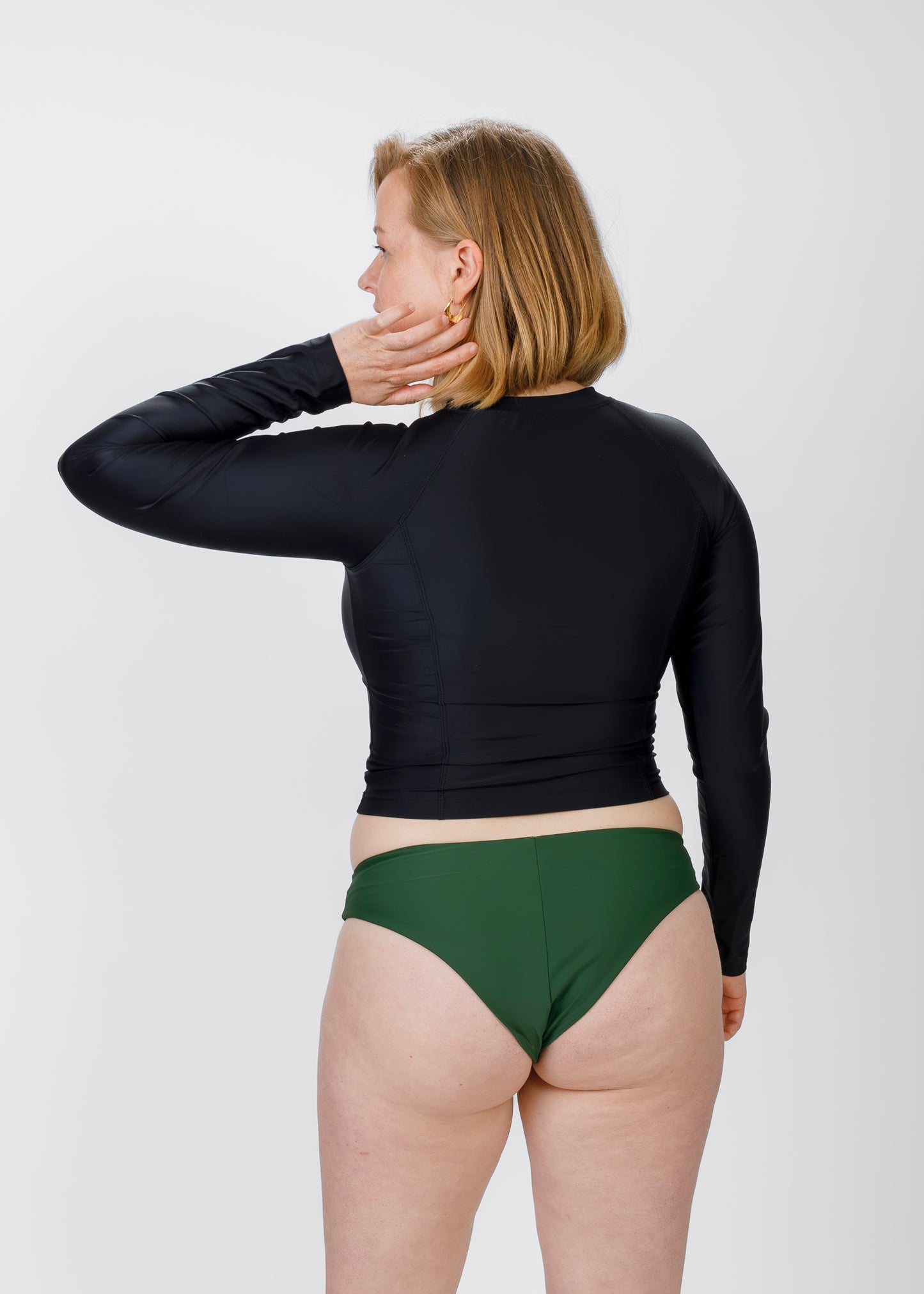 Side-back view of a blonde woman wearing Tamara’s black, long-sleeve rash guard and green bikini briefs, with one hand touching her hair against a white background.