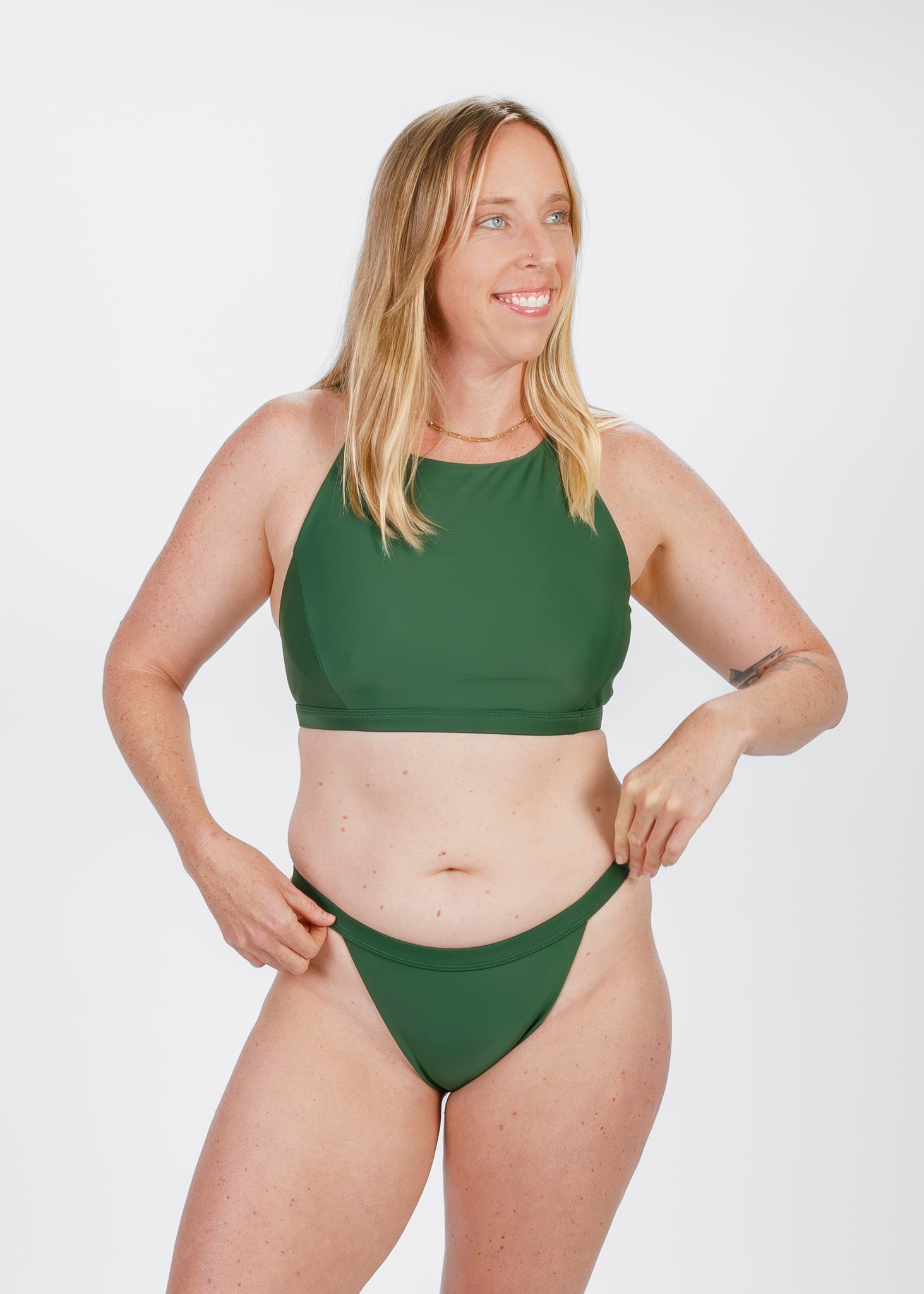 Model smiling and adjusting the waistband of her green high-waisted bikini, captured in a studio with a relaxed, confident expression.