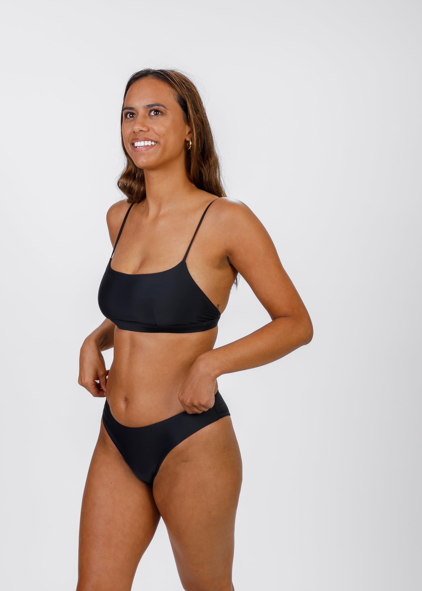 Woman in a black, thin-strapped bikini top and matching bottoms, smiling and adjusting the band at her waist against a white background.