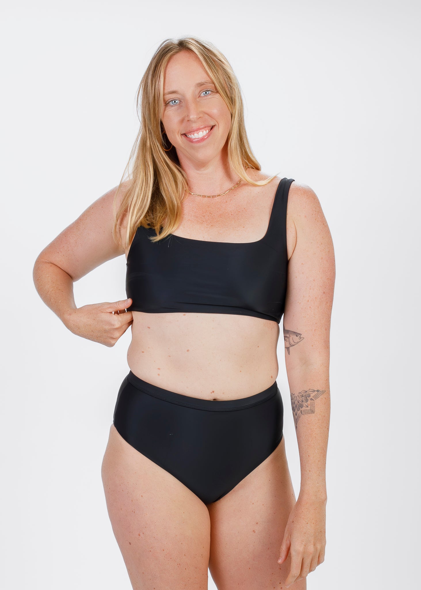 Smiling blonde woman wearing a black square-neck bikini top with wide straps, lightly pulling the underbust band while standing in high-waisted bottoms.