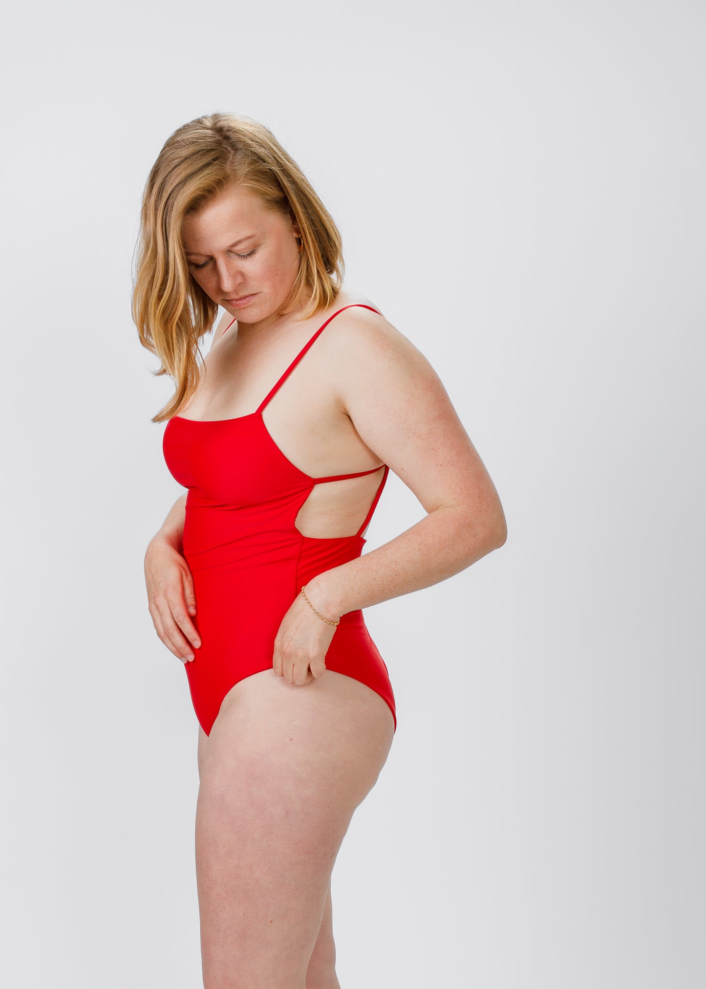 Side view of a blonde woman in a red, square-neck swimsuit with open side cut-outs, looking down as she adjusts the waist.