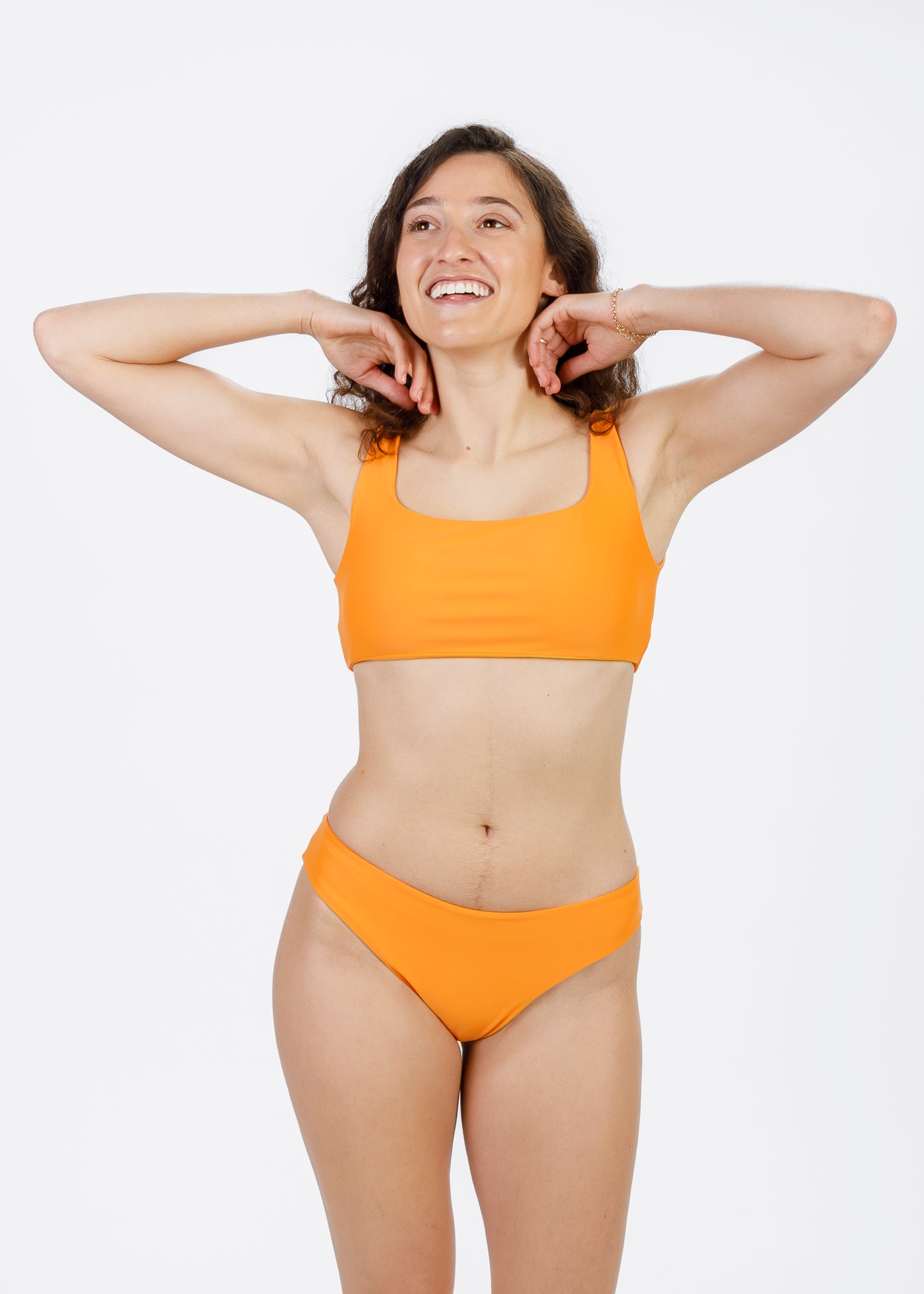 Woman with curly hair wearing a bright orange square-neck bikini top and matching bottoms, lifting her arms behind her head while smiling.
