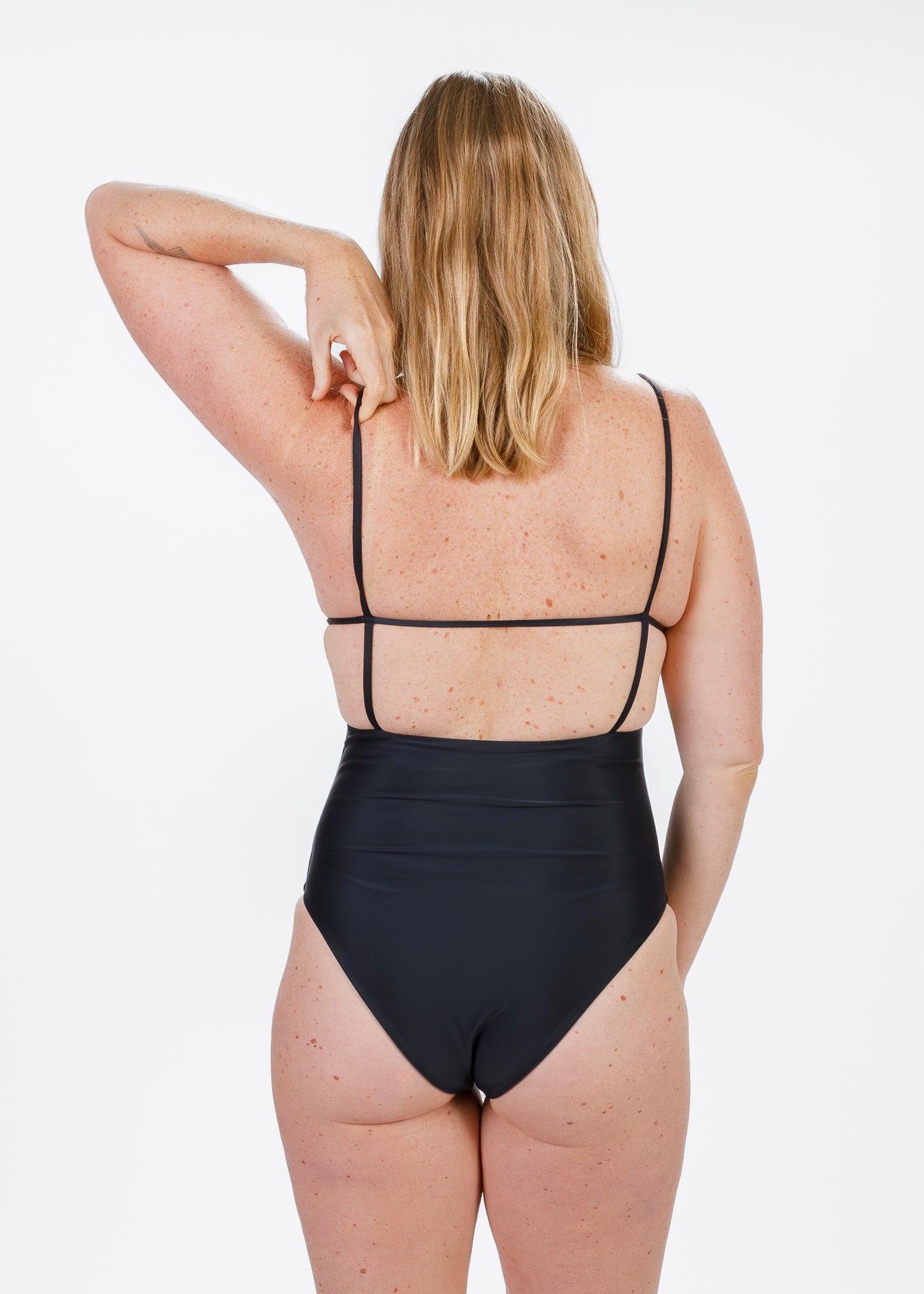 Rear view of a blonde woman in a black one-piece swimsuit, reaching over her shoulder to adjust the thin strap, highlighting the open back.