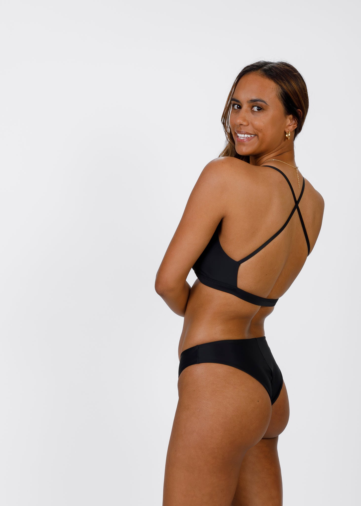 A woman with medium-brown skin looks over her shoulder while wearing a black bikini top with crisscross straps in the back. She smiles, emphasizing the open-back design and comfortable coverage of the bottoms.