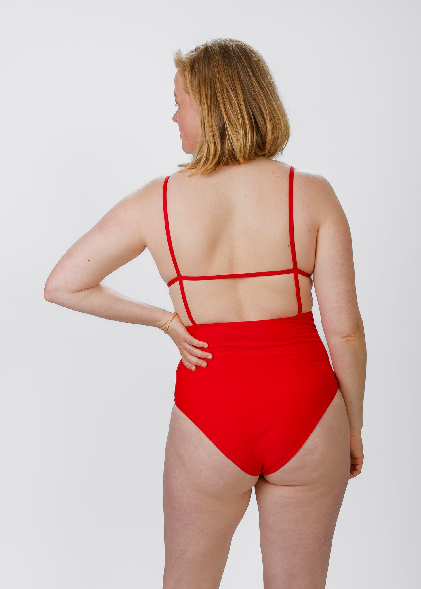 Blonde woman shown from behind in a red, thin-strapped one-piece swimsuit, highlighting the open back and high-waisted design.