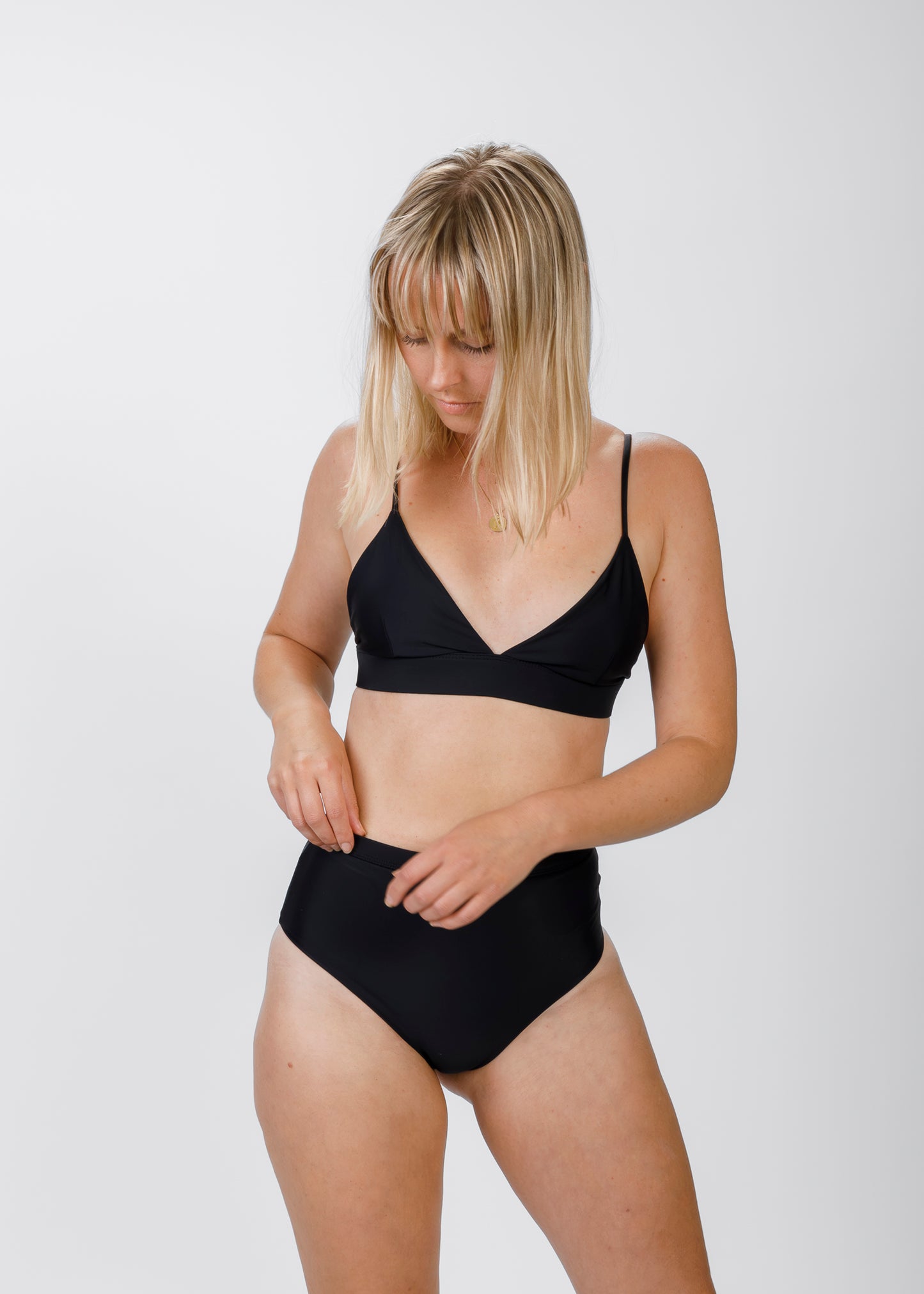 A blonde woman looks down as she adjusts her high-waisted bikini bottoms, drawing attention to the plunging neckline of her black triangle top. The slender straps and supportive underband highlight the minimalist, comfortable fit.