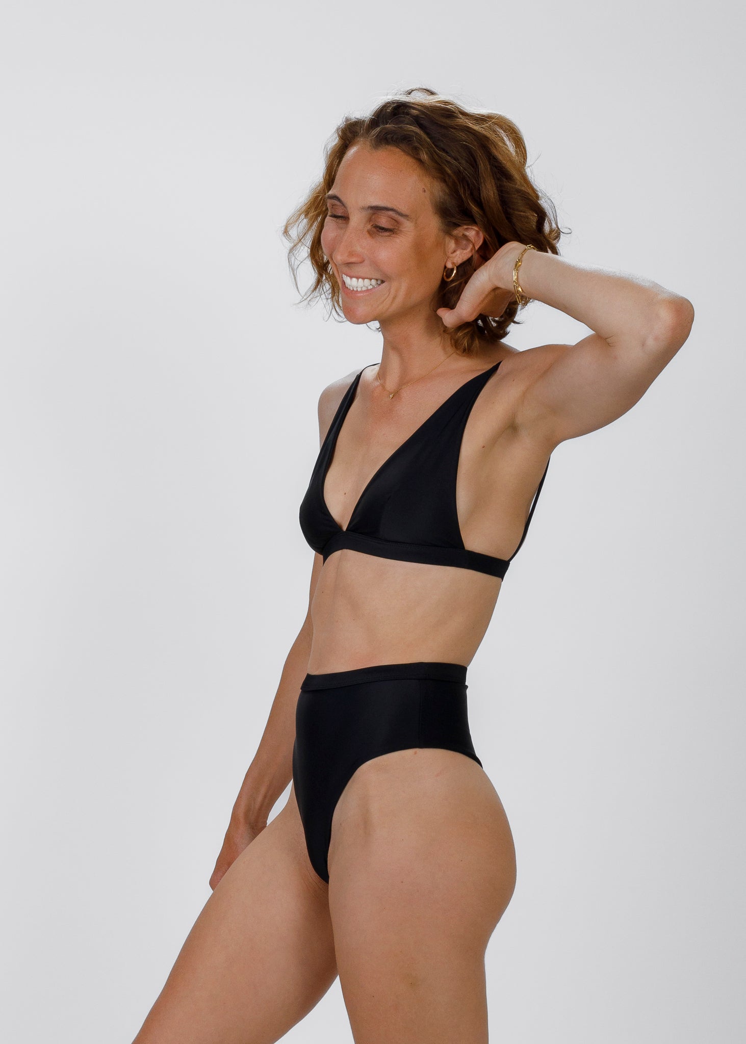 The same woman in a black bikini stands at a three-quarter angle, gently touching her hair. She pairs a deep V triangle top with high-waisted bottoms, showcasing a timeless, versatile silhouette.