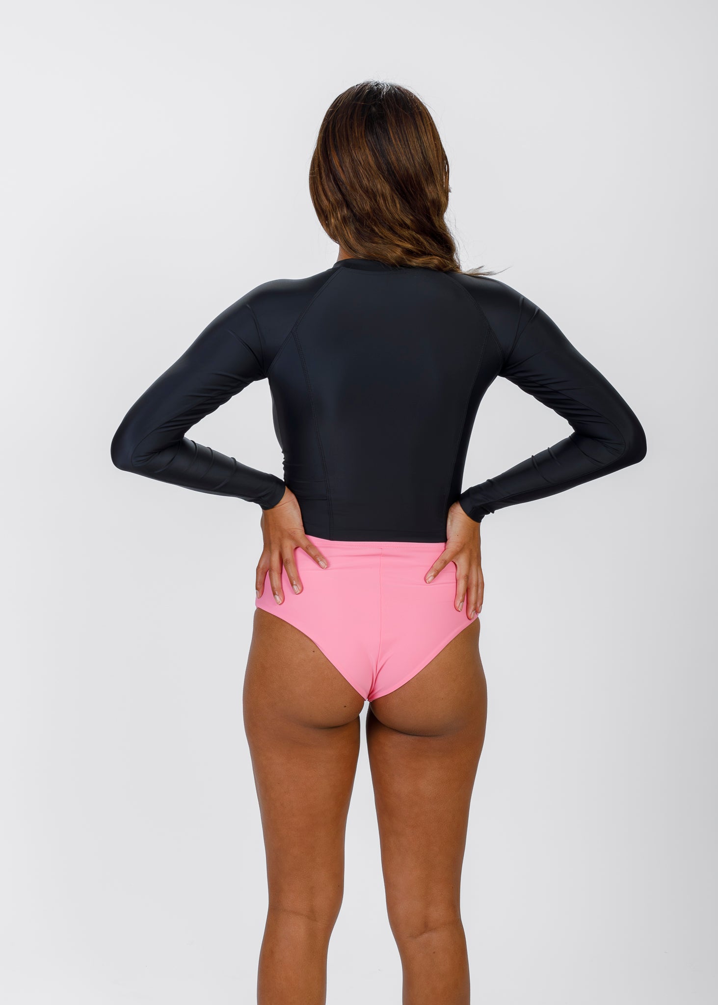 Back view of a woman in a black Tamara rash guard and pink high-waisted bikini bottoms, hands on her hips, facing away from the camera.