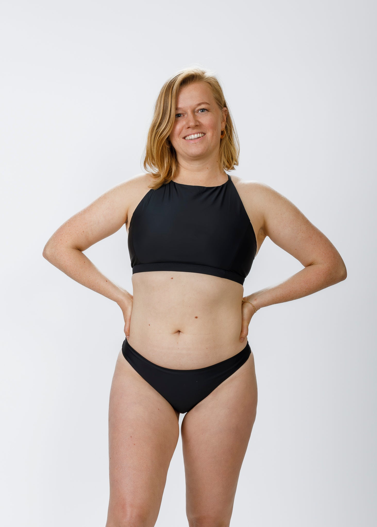 The same woman stands facing the camera in a black halter-style top paired with moderate-coverage bottoms. She places her hands on her hips, highlighting the bottoms’ secure waistband and timeless, flattering silhouette.