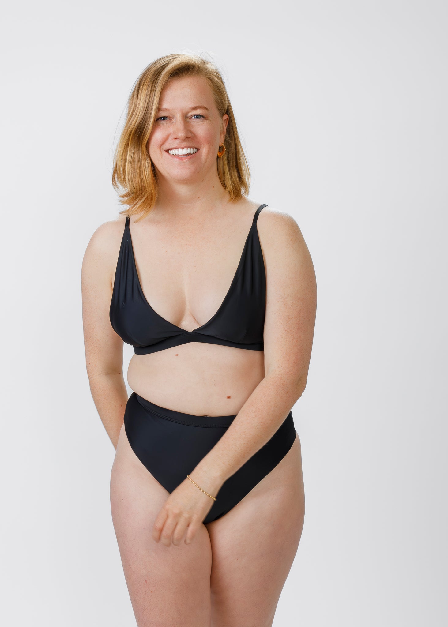 A blonde woman faces forward in a plunging black bikini top paired with high-waisted bottoms. She smiles confidently, showcasing the comfortable fit and timeless design of the bottoms.