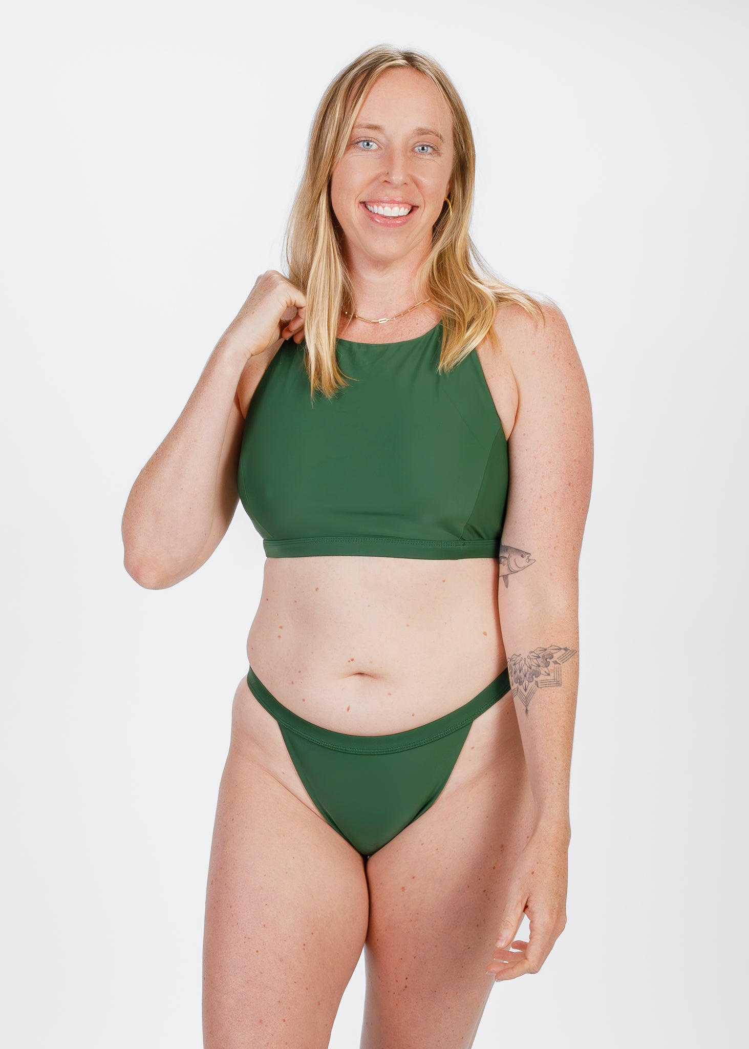 The same woman stands facing forward in a forest green high-neck bikini top and matching bottom. She smiles confidently, revealing subtle tattoos on her arms and reflecting the brand’s inclusive approach to swimwear.