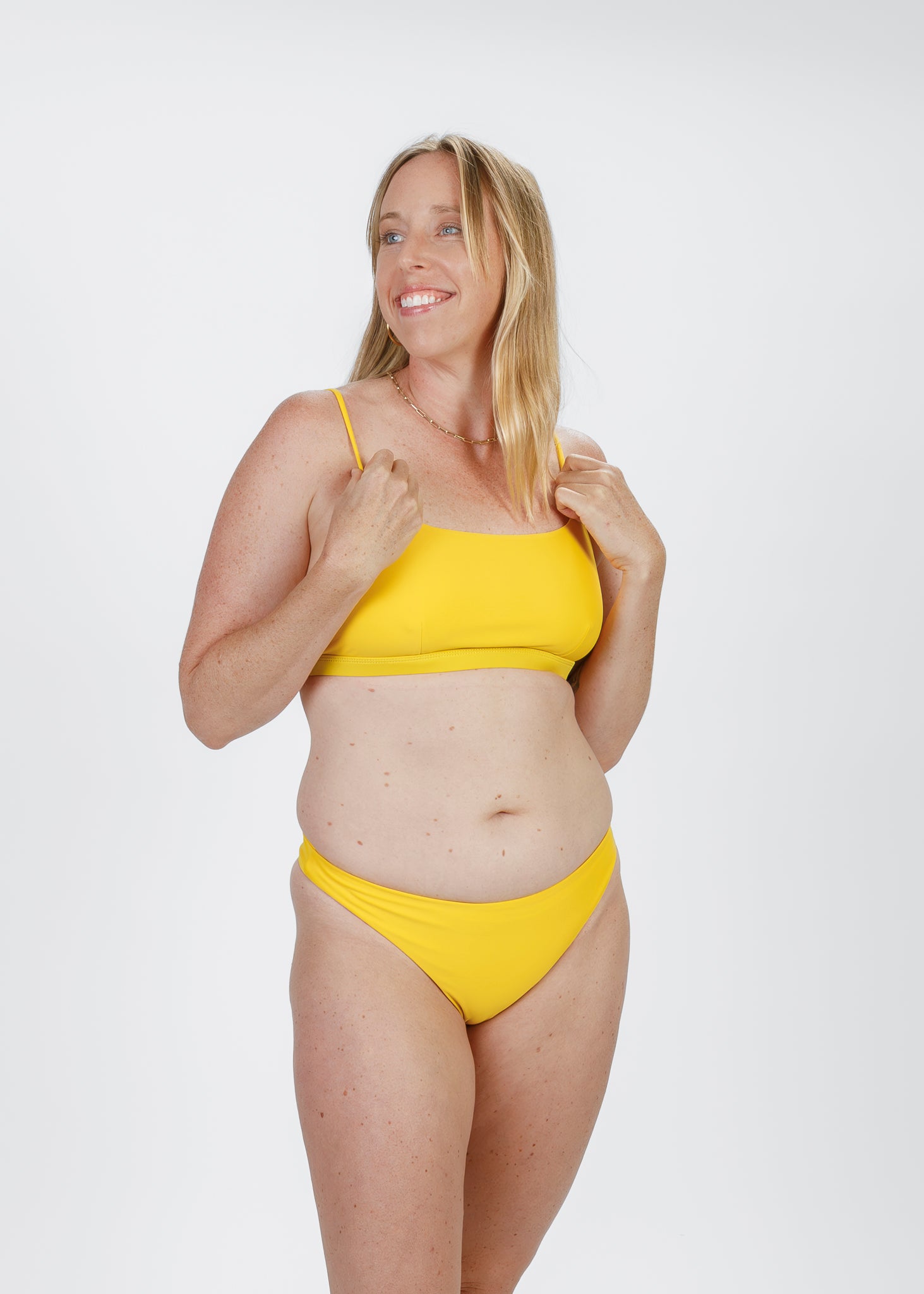 A blonde woman smiles while holding the straps of her yellow scoop-neck bikini top. She wears matching minimal-coverage bottoms that sit comfortably on her hips, highlighting a bright, cheerful swim look.