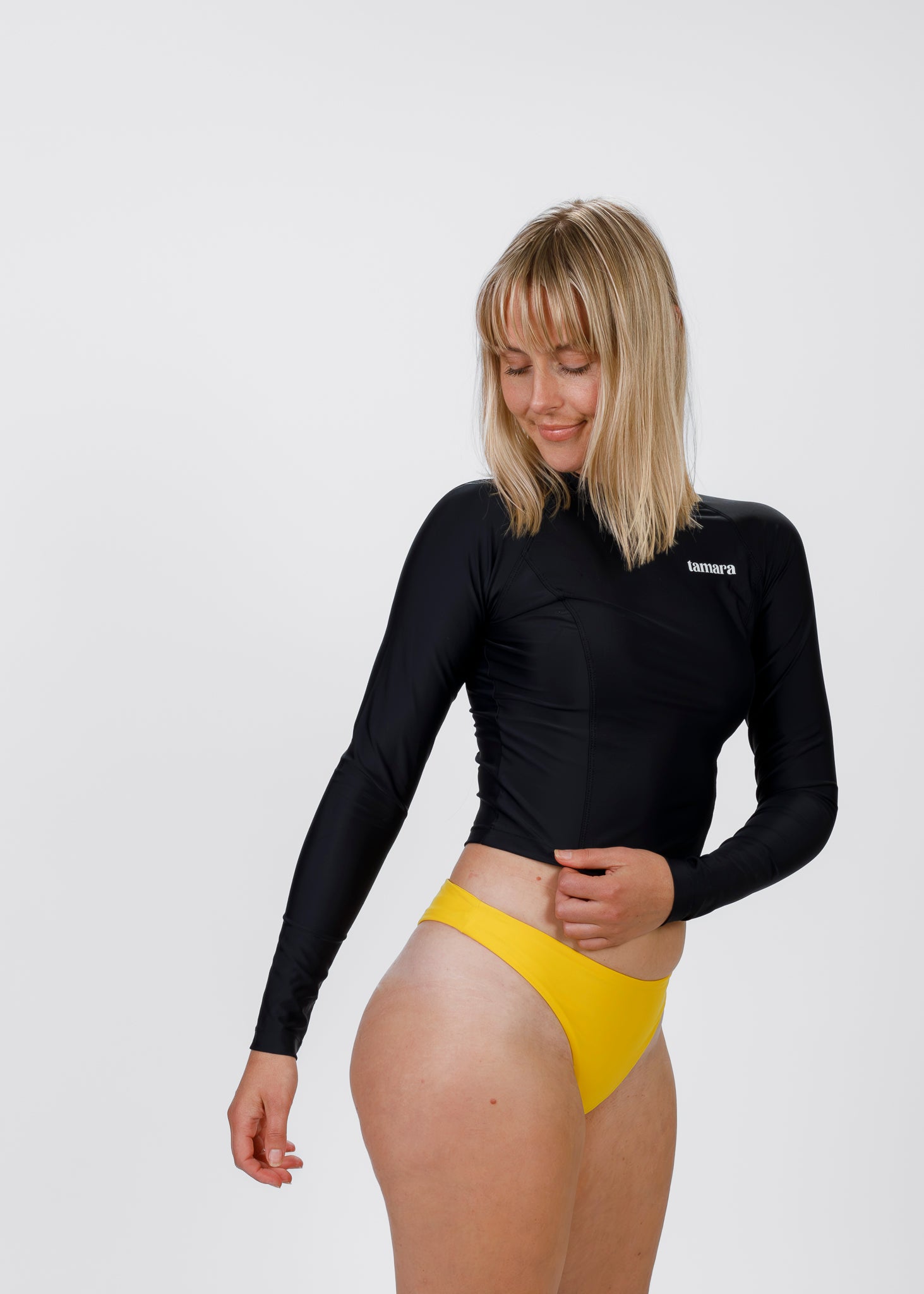 Blonde woman in Tamara’s black crop rash guard and yellow bikini bottoms, smiling softly while looking down against a white backdrop.