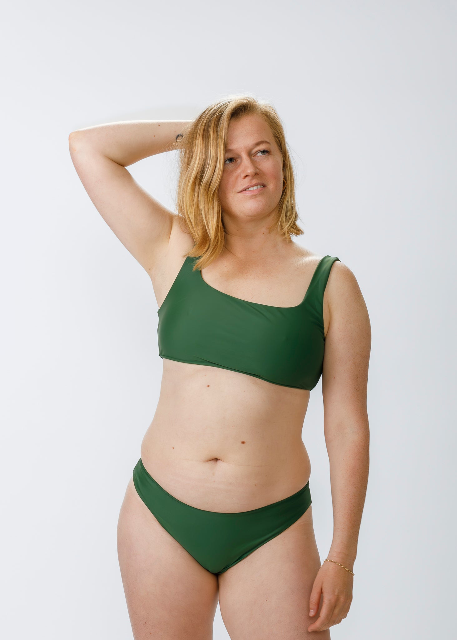 Blonde woman with shoulder-length hair wearing a green two-piece bikini, resting an arm behind her head against a plain white background.