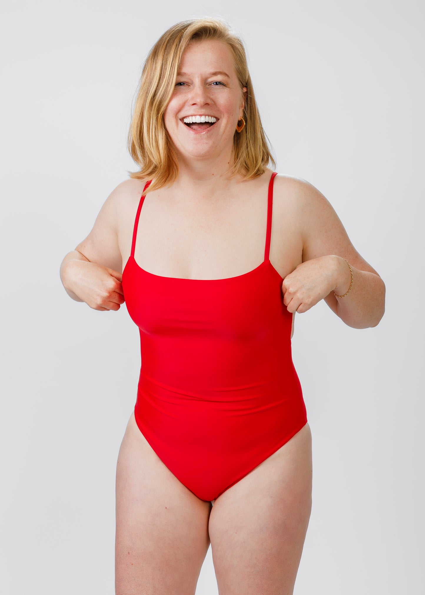 Woman with shoulder-length blonde hair wearing a bright red, thin-strapped one-piece swimsuit, laughing while adjusting the bust area.