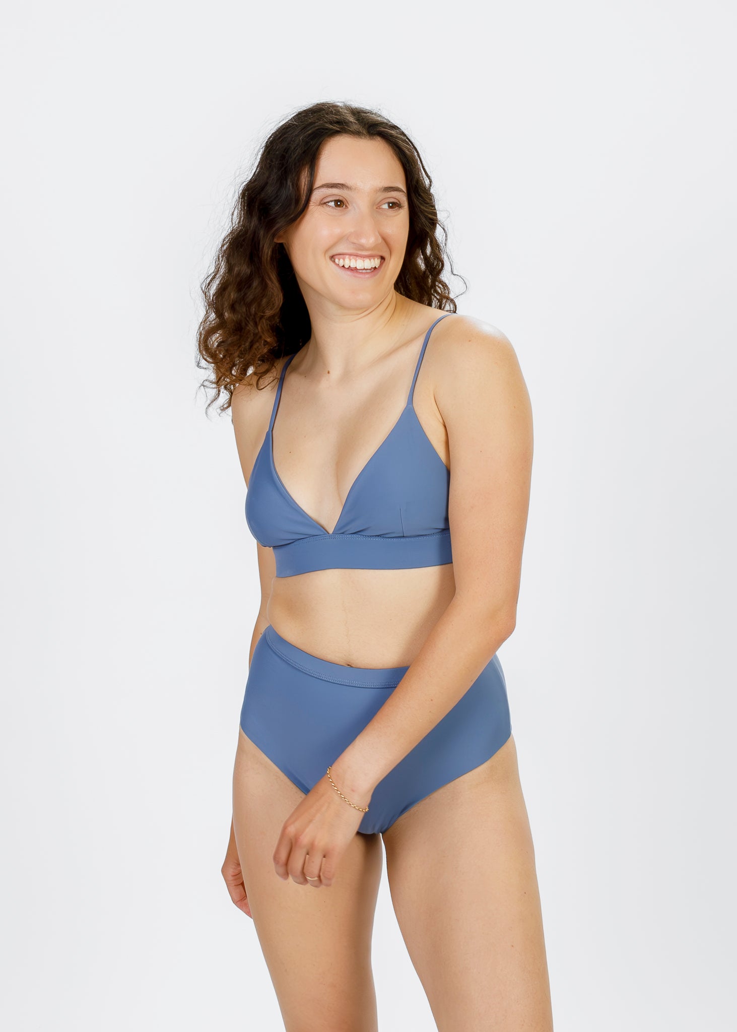 A brunette woman stands at a slight angle, wearing a light blue triangle bikini top with narrow straps and a wide underband. She smiles as she looks off-camera, showcasing the gentle scoop neckline and relaxed, supportive fit.