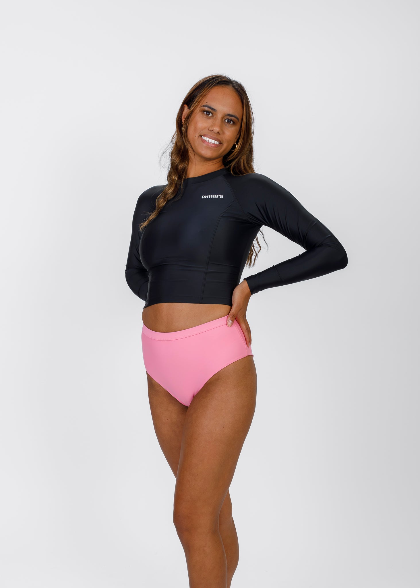 Woman with long, wavy hair wearing a black Tamara crop rash guard and pink high-waisted bikini bottoms, standing with one hand on her hip against a white backdrop.