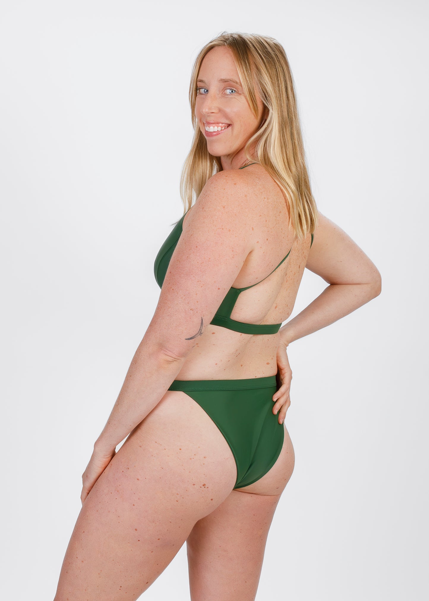 A fair-skinned woman with freckles turns slightly to reveal the back strap of her forest green halter bikini. She smiles, highlighting the supportive design and moderate coverage that align with the brand’s focus on comfort.